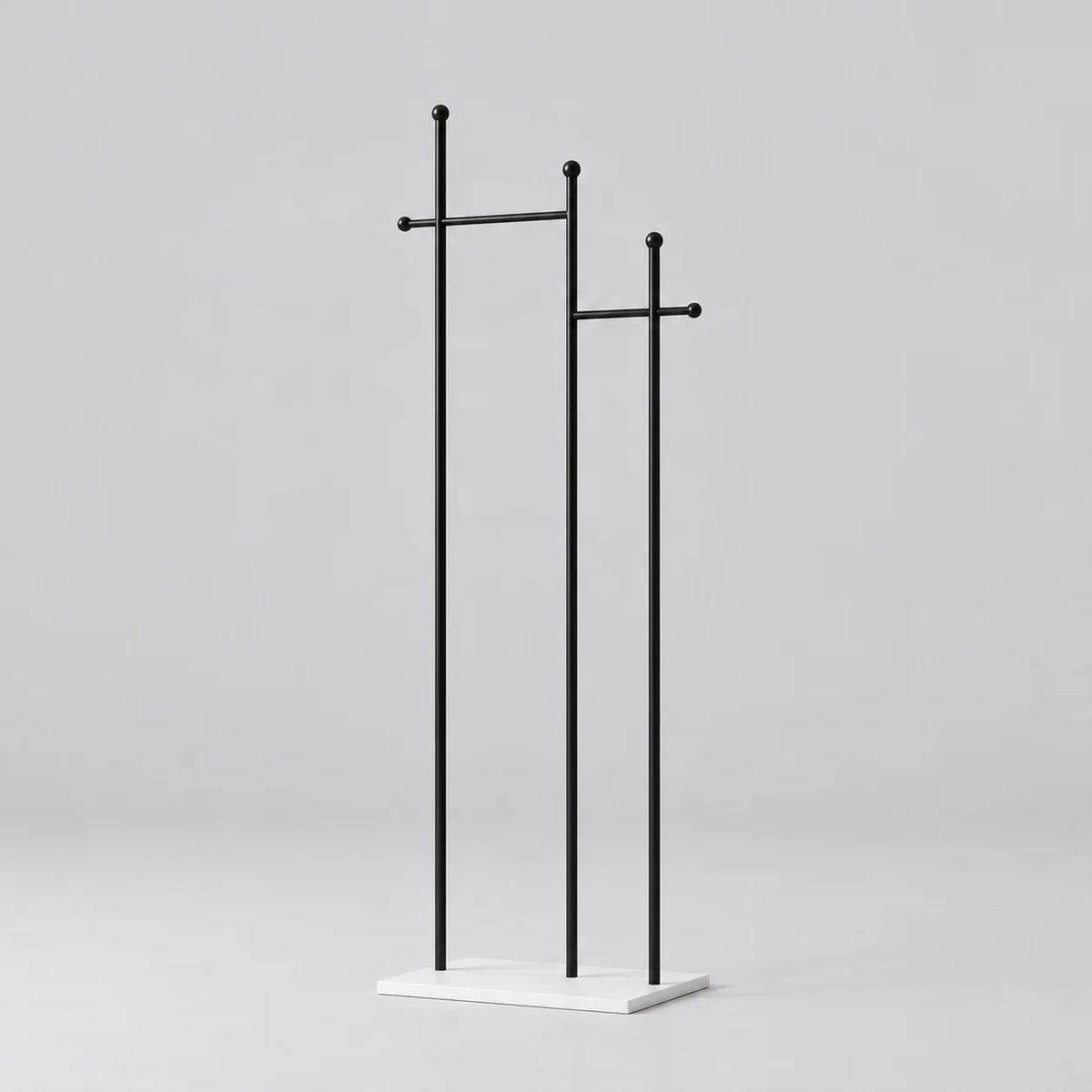 Porte-manteau sur pied à trois montants noirs de hauteurs décalées, deux traverses à embouts ronds et base rectangulaire blanche, sur fond gris