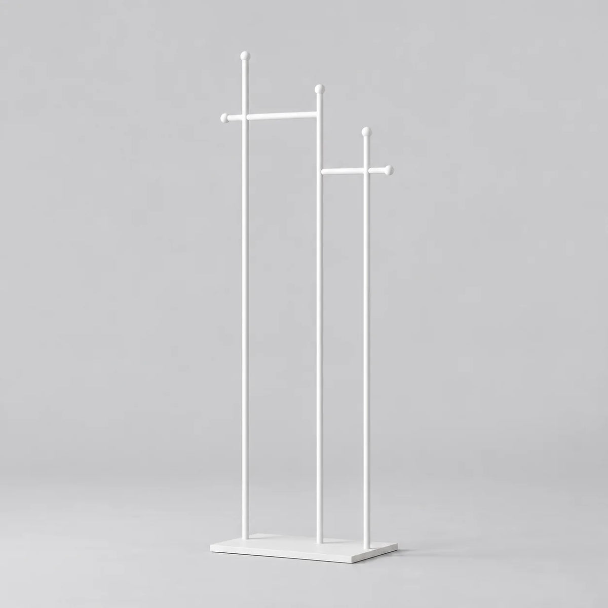 Porte-manteau sur pied blanc en métal, trois montants de hauteurs décalées, deux traverses à embouts ronds et base rectangulaire blanche, sur fond gris