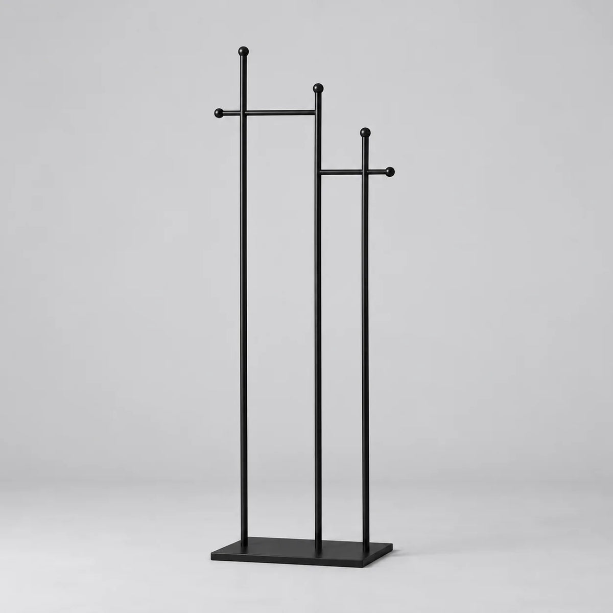 Porte-manteau sur pied en métal noir, trois montants de hauteurs décalées, traverses à embouts ronds et base rectangulaire, sur fond gris