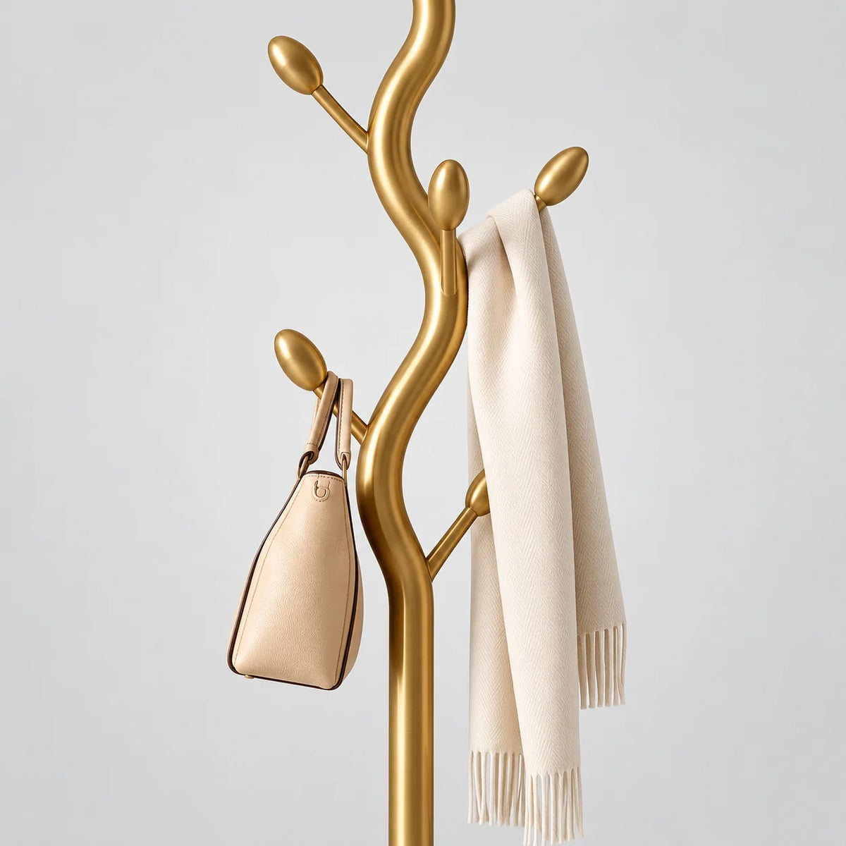 Détail des branches en acier doré brossé, du petit sac beige et de l’écharpe ivoire à franges sur fond gris