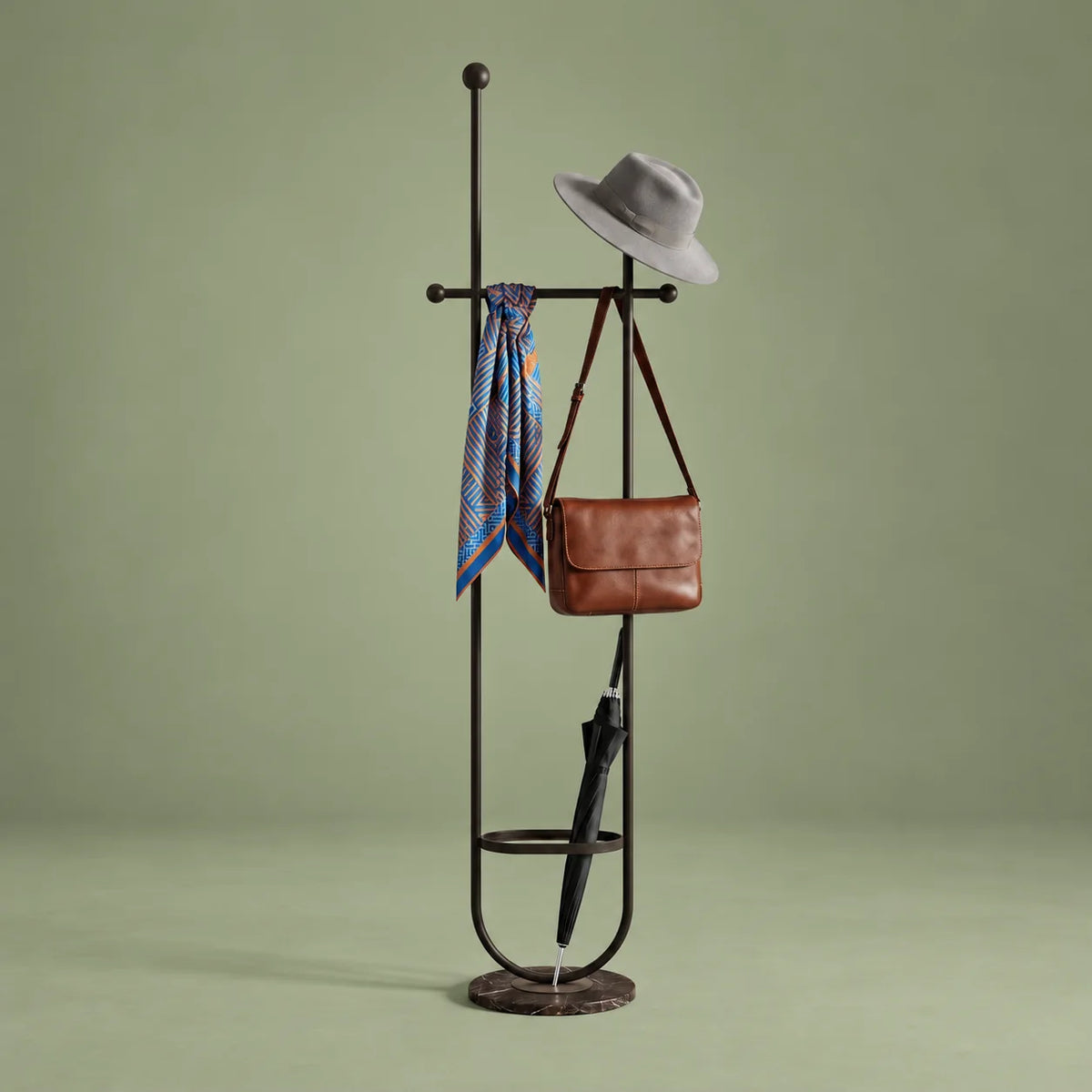 Porte-manteau sur pied en métal noir avec chapeau, foulard, sac et parapluie, anneau porte-parapluie et base ronde sombre sur fond vert sauge