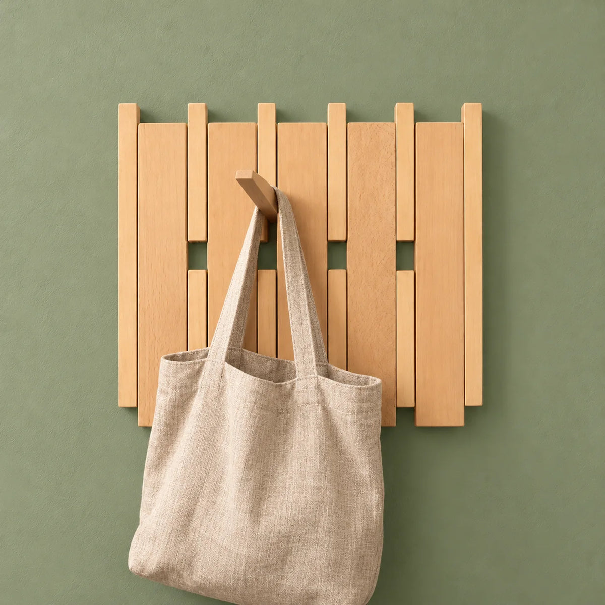 Porte-manteau carré en bois clair avec un sac en tissu suspendu sur fond vert sauge