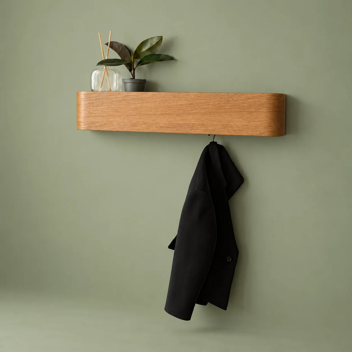 Porte-manteau mural en bois clair avec plante, diffuseur et manteau noir sur fond vert sauge