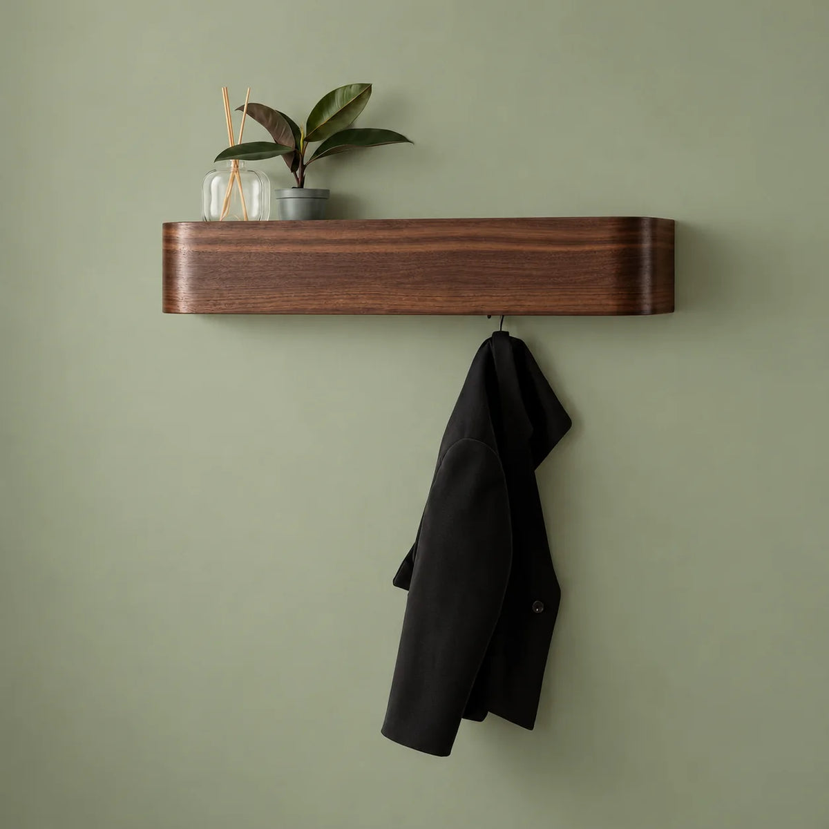Porte-manteau mural en bois foncé avec plante, diffuseur et manteau noir sur fond vert sauge