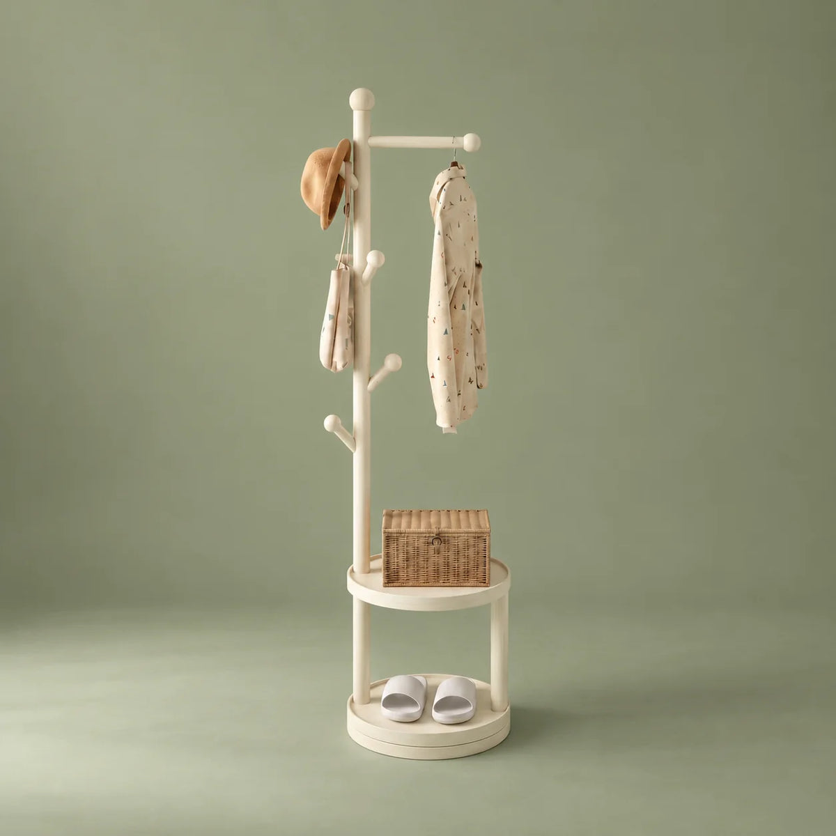 Porte-manteau sur pied en bois blanc avec patères, deux étagères rondes, chapeau, sac, vêtement, panier et chaussures sur fond vert sauge