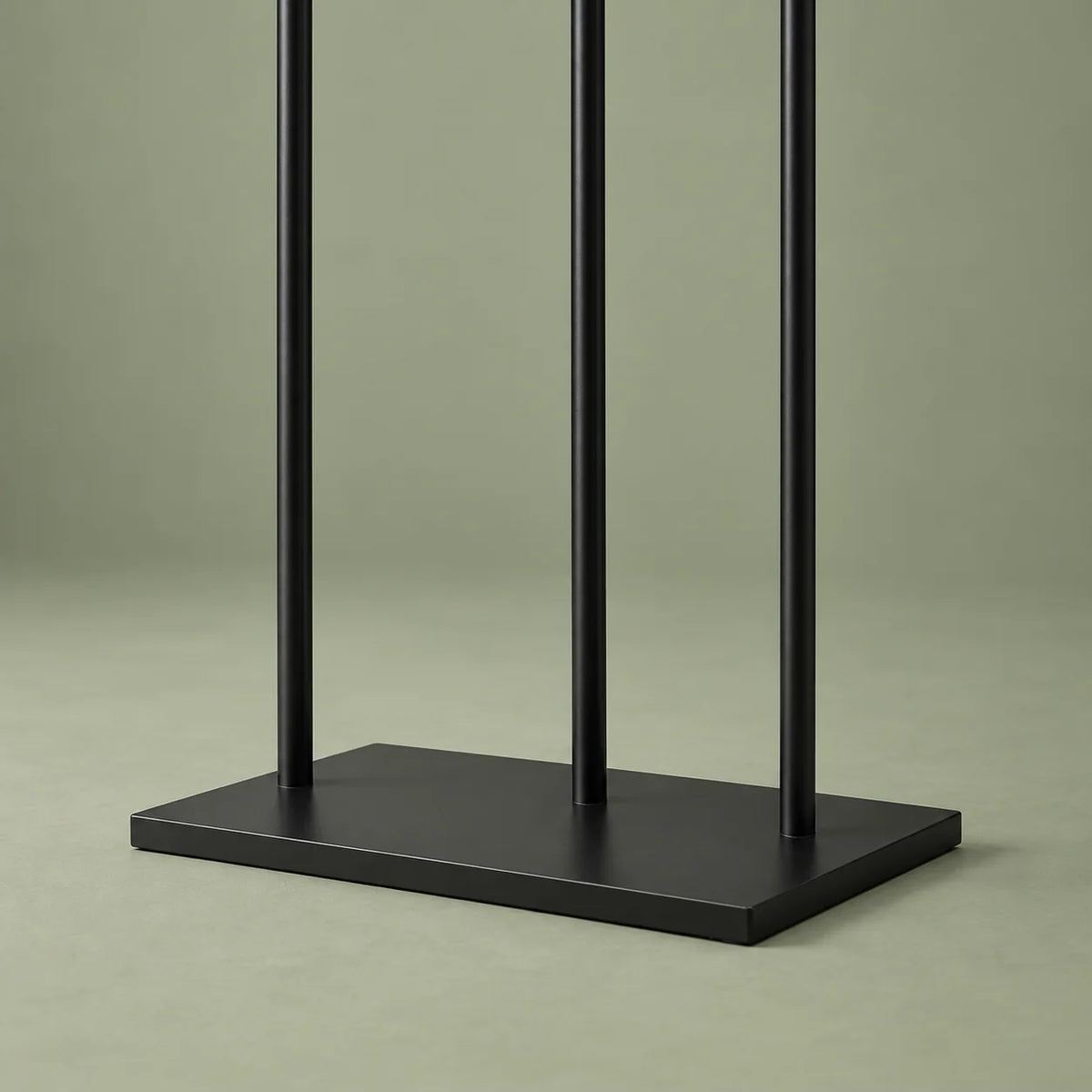 Gros plan sur la base rectangulaire noire et les trois montants métalliques du porte-manteau, sur fond vert sauge